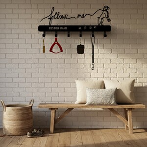 Pode incluir: Suporte de trela para cães montado na parede de metal preto com as palavras "follow me" e "CUSTOM NAME". Apresenta ganchos para trelas e acessórios, uma silhueta de cão e pegadas. Inclui uma coleira vermelha, uma bolsa preta e uma trela preta.