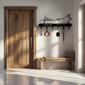Op de afbeelding: Een zwarte metalen hondenriemhouder met de woorden "follow me" en "CUSTOM NAME". Het heeft haken voor riemen en accessoires, met pootafdrukken en een honden silhouet. Inclusief een rood tuigje en zwarte accessoires.