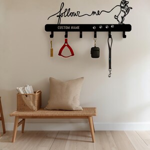 Op de afbeelding: Zwarte metalen hondenriemhouder met de woorden "follow me" en "CUSTOM NAME". Voorzien van haken voor riemen en accessoires. Een rood hondentuigje, een zwarte tas en een zwarte riem hangen aan de haken. Honden silhouet rechts.