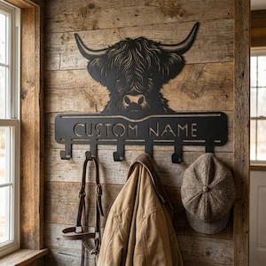 Op de afbeelding: Zwarte metalen wandkapstok met een Highland koeienontwerp. De kapstok heeft haken om items op te hangen en de tekst "CUSTOM NAME". Een bruine jas, een pet en een hoofdstel hangen aan de haken.