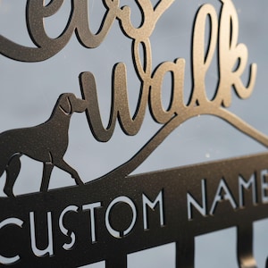 May include: A black metal sign with the words "Let's Walk" in a cursive font, featuring a dog silhouette. Below the text is a line with the words "CUSTOM NAME" in capital letters. The sign has a matte finish.