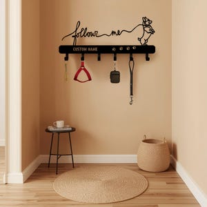 Puede incluir: Soporte de correa para perros de metal negro con las palabras "follow me" y "CUSTOM NAME". El soporte presenta una silueta de perro, huellas de patas y ganchos para correas y accesorios. Un arnés rojo para perros, una funda negra y una correa negra cuelgan de los ganchos.