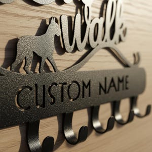 May include: A black metal wall-mounted dog leash holder with the words "Walk" and "Custom Name" cut out. A silhouette of a dog is above the name plate. Includes several hooks for leashes and accessories. Mounted on a light wood background.