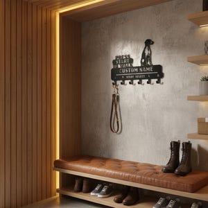 May include: A black metal dog leash holder with the text "PRESS START TO WALK CUSTOM NAME". The holder features a silhouette of a dog and five hooks. A brown leather leash hangs from one hook. The holder is mounted on a gray wall.
