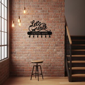 Puede incluir: Soporte de correa para perros de metal negro con las palabras "Let's Walk" y una silueta de perro. Debajo del texto hay ganchos para correas y las palabras "CUSTOM NAME". El soporte está montado en una pared de ladrillo.