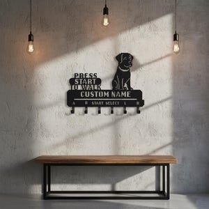 以下が含まれることがあります： 犬のシルエットと「PRESS START TO WALK CUSTOM NAME」の文字が入った黒い金属製のキーホルダー。鍵用のフックと下の木製ベンチが含まれています。デザインはテクスチャード加工された壁に取り付けられています。