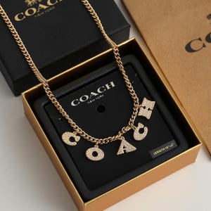 May include: Gold-toned necklace with charms spelling "COACH" in a gift box. The necklace has a chain design and the charms are encrusted with small, sparkling stones. The box is black with the brand name in white.
