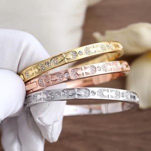 May include: Three gold, rose gold, and silver-toned bangles. Each bangle features a repeating pattern of the letter "C" and is embellished with small, sparkling crystals. The bangles are held by a gloved hand.