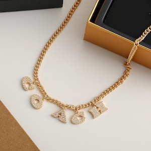 May include: Gold-toned chain necklace with dangling letter charms spelling out "OACH". Each letter is encrusted with small, sparkling crystals. The necklace is displayed on a white surface, with a gold box in the background.