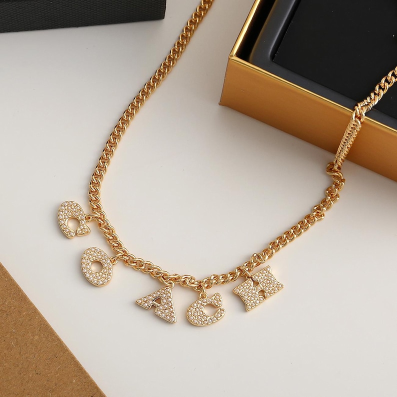 May include: Gold-toned chain necklace with dangling letter charms spelling out "OACH". Each letter is encrusted with small, sparkling crystals. The necklace is displayed on a white surface, with a gold box in the background.