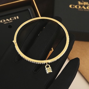 Op de afbeelding: Een goudkleurige armband met een kleine hangslotbedel. De armband is aan de bovenste helft bezet met kleine, heldere steentjes. De hangslotbedel is gedetailleerd met een herhalend patroon. De armband wordt op een zwarte ondergrond getoond.