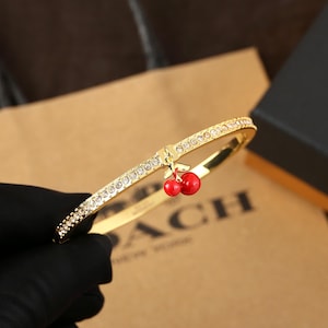 May include: Gold-tone bracelet with a cherry charm. The bracelet is adorned with sparkling crystals along the top and features a small, red cherry pendant. The bracelet is a stylish accessory.