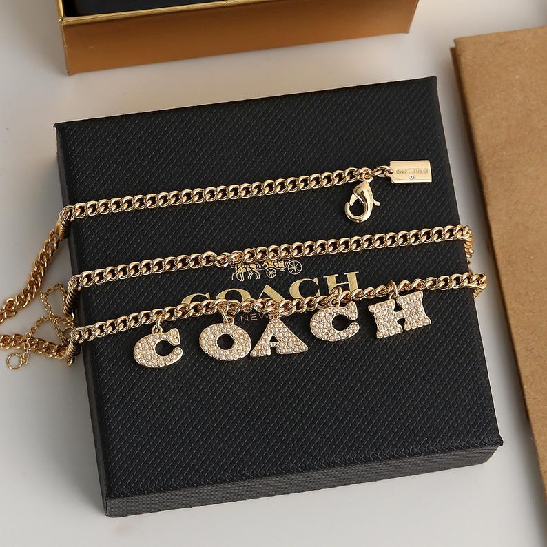 May include: A gold-colored necklace with the word "COACH" spelled out in individual letter charms. The necklace is on a black box with the Coach logo. The chain is a curb link style.