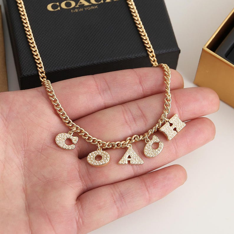 May include: Gold-toned necklace with a chain and multiple charms spelling out "COACH". The charms are encrusted with small, sparkling stones. The necklace is displayed on a hand, with a black box in the background.