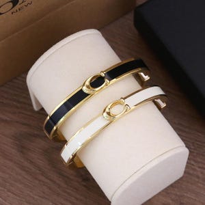 Op de afbeelding: Twee goudkleurige metalen armbanden, één zwart en één wit, worden tentoongesteld op een witte standaard. Elke armband heeft een goudkleurige sluiting met een cirkelvormig ontwerp. De zwarte en witte banden hebben een glanzende afwerking.