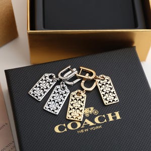 May include: Silver and gold tag earrings with the Coach logo pattern. The earrings are displayed on a black textured surface with the Coach New York logo in gold. A gold box is in the background.