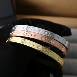 May include: Three bangles in silver, rose gold, and gold. Each features a repeating logo pattern and sparkling crystal accents. These stylish bracelets are a fashionable accessory.
