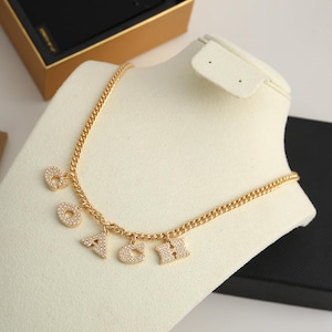 May include: Gold-toned necklace with the letters COACH spelled out in sparkling, clear gemstones. The necklace has a curb chain and is displayed on a white jewelry bust. The letters are dangling from the chain.