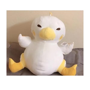 Könnte beinhalten: Ein großes, plüschiges weißes Vogelspielzeug mit gelbem Schnabel, Füßen und Akzenten um die Augen. Das Stofftier hat ausgestreckte Flügel und sitzt auf einer grauen Oberfläche. Der Vogel hat kleine braune Augen und einen kleinen gelben Kamm.