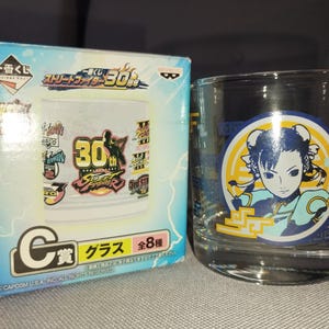 Op de afbeelding: Een helder glas met een Street Fighter 30th Anniversary-ontwerp. Het glas heeft een cirkelvormige afbeelding van een personage in een blauw en geel kostuum. Een doos met Japanse tekst en Street Fighter-graphics staat naast het glas.