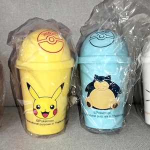 May include: Four colorful Pokemon-themed plastic cups. Each cup features a different character: Jigglypuff, Pikachu, Snorlax, and Meowth. The cups are in pink, yellow, blue, and white, and are sealed in plastic.