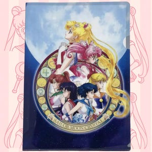 Puede incluir: Un archivador de plástico transparente con una colorida ilustración de personajes de Sailor Moon Crystal. La obra presenta múltiples figuras en varias poses, sobre un fondo de cielo azul y luna, con el texto "SAILOR MOON CRYSTAL".