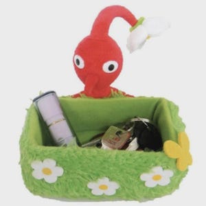 Puede incluir: Una cesta de almacenamiento verde de felpa con un personaje Pikmin rojo asomándose por el borde. La cesta está decorada con margaritas blancas y una mariposa amarilla. Dentro hay llaves y un tubo pequeño. El Pikmin tiene una flor blanca en la cabeza.