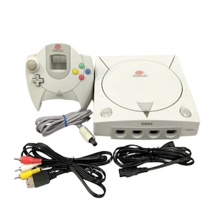 May include: A white Sega Dreamcast video game console with a matching controller, power cord, and AV cables. The console has the Dreamcast logo and the word "SEGA" printed on the front. The controller has a directional pad and colored action buttons.