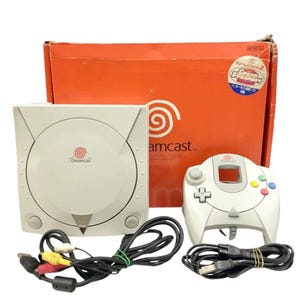Puede incluir: Una consola de videojuegos Dreamcast blanca con un mando a juego, cables y una caja naranja. La consola y el mando tienen el logotipo de Dreamcast. La caja tiene texto japonés y una pegatina.