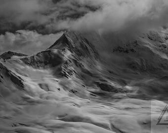 May include: Black and white landscape photograph of a snow-covered mountain range under a cloudy sky. The image captures the rugged terrain and the interplay of light and shadow across the peaks and valleys. The photograph is signed by Edge Photography.