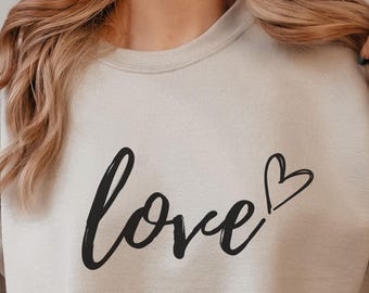 May include: Beige sweatshirt with the word "love" in black script and a heart symbol. The sweatshirt is a neutral color and has a crew neck. A white mug is held in the hand.