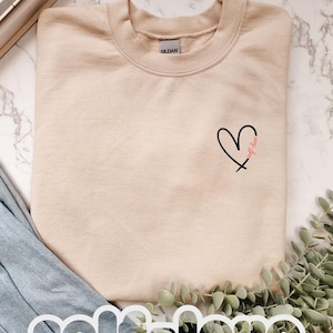 Könnte beinhalten: Beiges Sweatshirt mit schwarzem Herz-Grafik und dem Schriftzug "self-love" in roter Schrift. Das Sweatshirt liegt auf einer Marmoroberfläche, im Hintergrund Denim und Grün. Das Wort "self-love" ist auch unten in Weiß aufgedruckt.