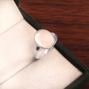 Peut inclure: Une bague en argent avec une grosse pierre précieuse ovale rose pâle. La pierre est sertie dans un anneau en argent poli. La bague est présentée dans un écrin vert foncé avec un intérieur blanc. C'est un bijou.