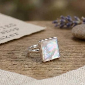 May include: A silver ring featuring a square-shaped iridescent mother of pearl inlay. The polished metal band supports a flat, reflective stone surface that displays shifting hues of pink, blue, and white under the light, creating a subtle glow.