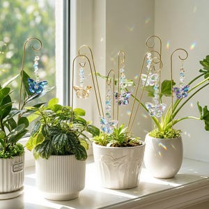 Può includere: Una collezione di supporti per piante in metallo dorato, ognuno con una farfalla di cristallo pendente in colori blu, ambra, viola e trasparente. I supporti sono inseriti nei vasi e presentano perline sfaccettate che riflettono la luce.