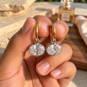 May include: A pair of earrings featuring polished gold-tone hoop tops connected to oval-shaped, clear faceted stones set in silver-tone metal prongs. The design combines a metallic gold hoop with a dangling, brilliant-cut oval stone setting.