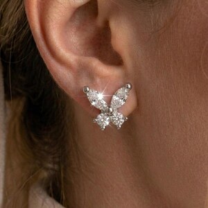 May include: A silver-toned stud earring shaped like a butterfly, featuring two marquise-cut clear stones for the upper wings and two round-cut clear stones for the lower wings, all set in a metallic frame that catches the light with a bright sparkle.