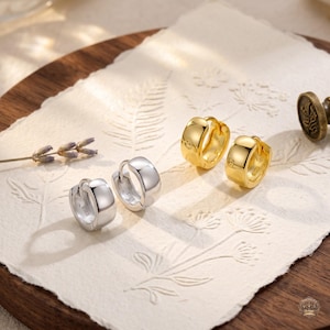 May include: A pair of silver-toned hoop earrings and a pair of gold-toned hoop earrings. Each earring features a smooth, polished finish with a hinged closure mechanism. The earrings are small, circular, and have a thick, rounded profile design.
