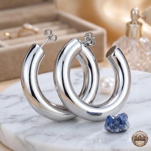 May include: A pair of thick, polished silver-tone hoop earrings with a smooth, rounded profile. The earrings feature a standard post-back closure mechanism. The metal surface is highly reflective, showing clear highlights and a bright metallic finish.