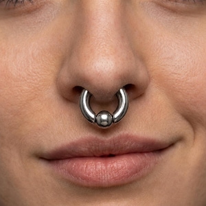 May include: A circular septum ring made of polished silver-toned metal. The jewelry features a smooth, continuous hoop design with a single, central spherical bead that sits securely between the two ends of the ring, creating a seamless look.