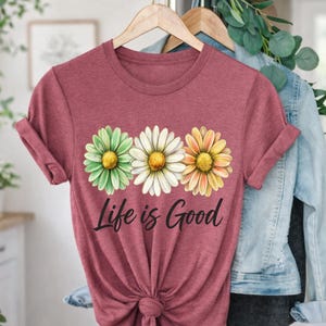 May include: A maroon t-shirt with three daisy flower graphics in green, white, and yellow. The text "Life is Good" is printed below the flowers in a black script font. The shirt is hanging on a wooden hanger.