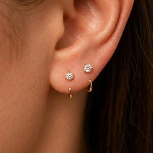 May include: Two gold stud earrings featuring round clear stones in a prong setting, paired with two small gold hoop earrings. The jewelry is arranged in a stacked configuration on the earlobe, showcasing a polished metallic finish and clear gems.