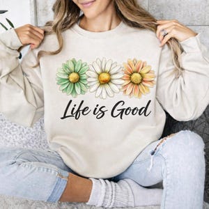 May include: Beige sweatshirt featuring three daisy flowers in green, white, and orange, with yellow centers. The phrase "Life is Good" is printed in black script below the flowers. The sweatshirt is a casual, comfortable style.