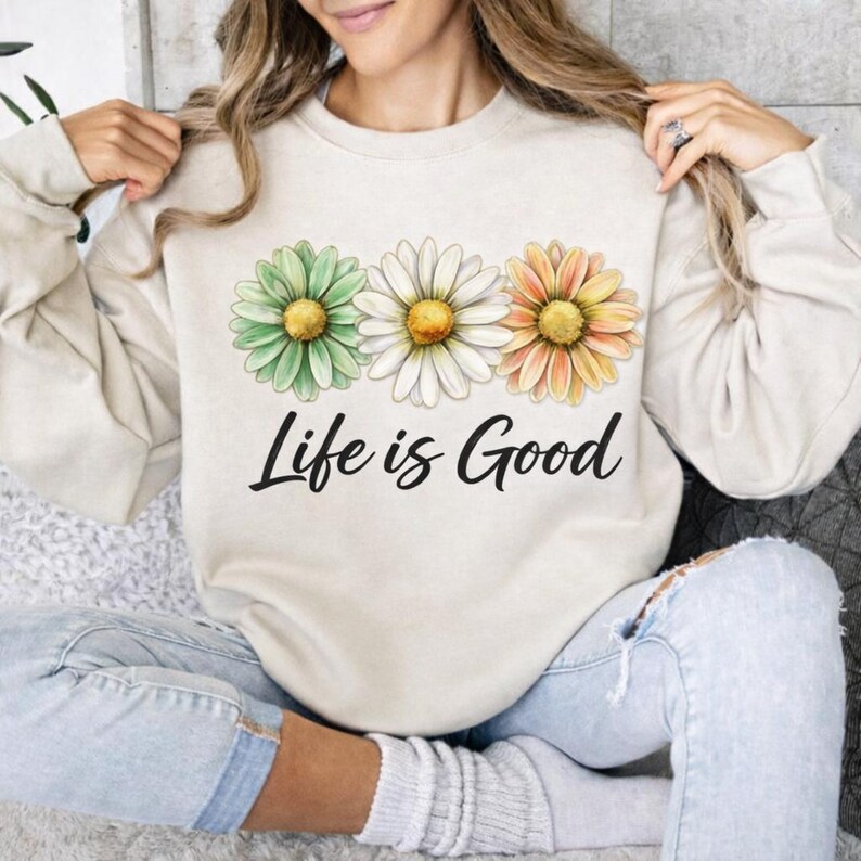 May include: Beige sweatshirt featuring three daisy flowers in green, white, and orange, with yellow centers. The phrase "Life is Good" is printed in black script below the flowers. The sweatshirt is a casual, comfortable style.