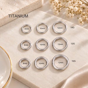 May include: A collection of silver-colored titanium circular rings arranged in a grid. The rings are labeled with sizes 8mm, 10mm, and 12mm, and gauges 14G, 12G, and 10G. Each ring features a seamless hinge closure design for body jewelry applications.