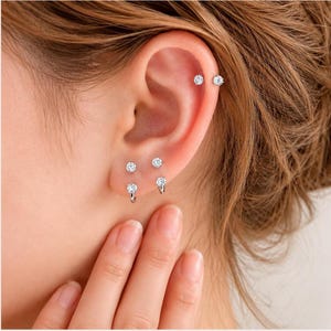 May include: Close-up of an ear with multiple piercings. The ear features three pairs of small, sparkling stud earrings and two small hoop earrings with a single, clear gem. The earrings are silver-toned and create a delicate, elegant look.