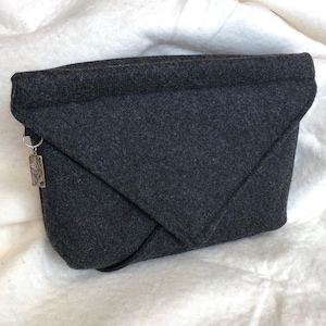 May include: A dark gray felt clutch bag with a folded design. The bag has a rectangular shape with a flap closure. A small charm hangs from a silver ring on the side of the bag.