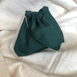 May include: A dark green drawstring pouch with a white interior and a white drawstring. The pouch is made of a soft fabric and is gathered at the top. The pouch is sitting on a white, textured surface.