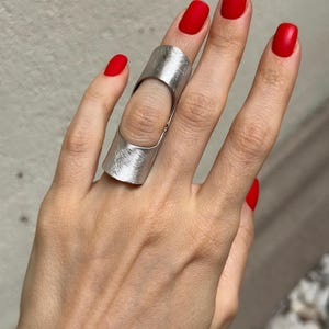 May include: A silver-toned ring with a textured surface, worn on a finger. The ring has a unique design that covers a portion of the finger. The hand has red nail polish.