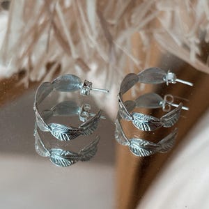 May include: A pair of silver hoop earrings. Each earring has a layered design of stylized leaves. The earrings are shown on a reflective surface, with soft, blurred background elements.